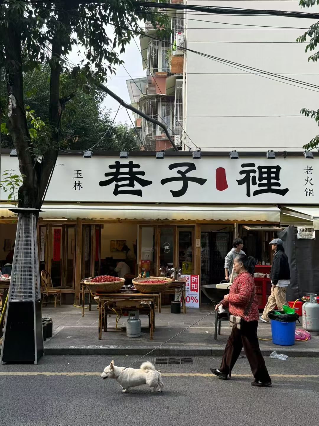 玉林「巷子裡」老火锅门头：白底黑字招牌，门口竹筐里晒着辣椒，街边有人遛狗